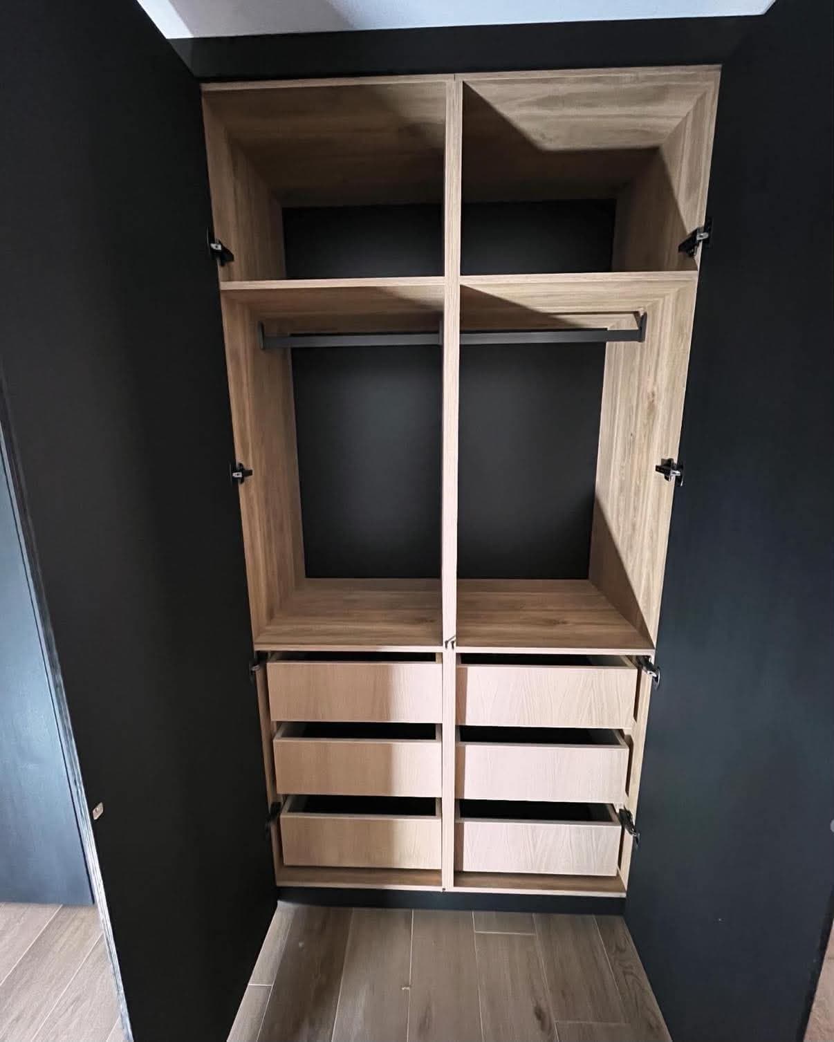 Intérieur de dressing en panneau décor chêne, avec penderie, étagères et six tiroirs, encastré dans une niche noire.