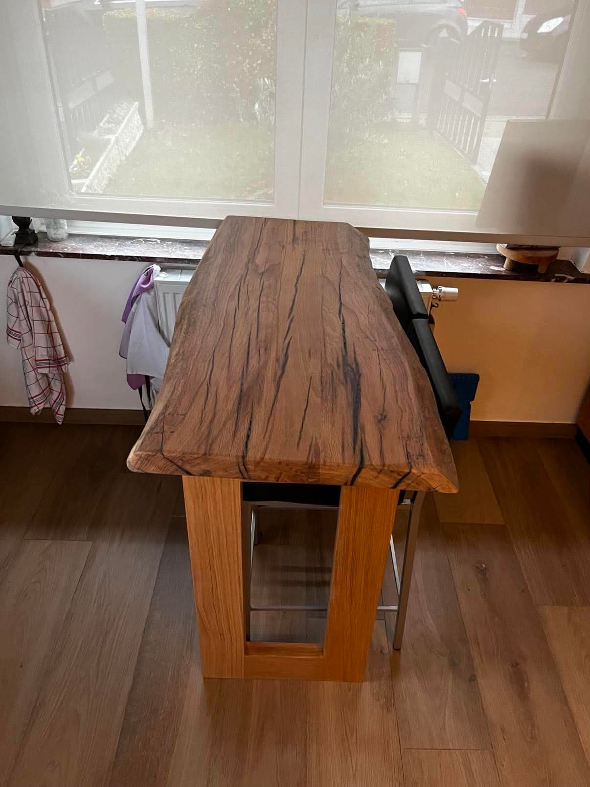 Table haute au plateau de chêne massif à bord naturel et fentes remplies de résine noire, posée sur un pied en panneau de chêne.
