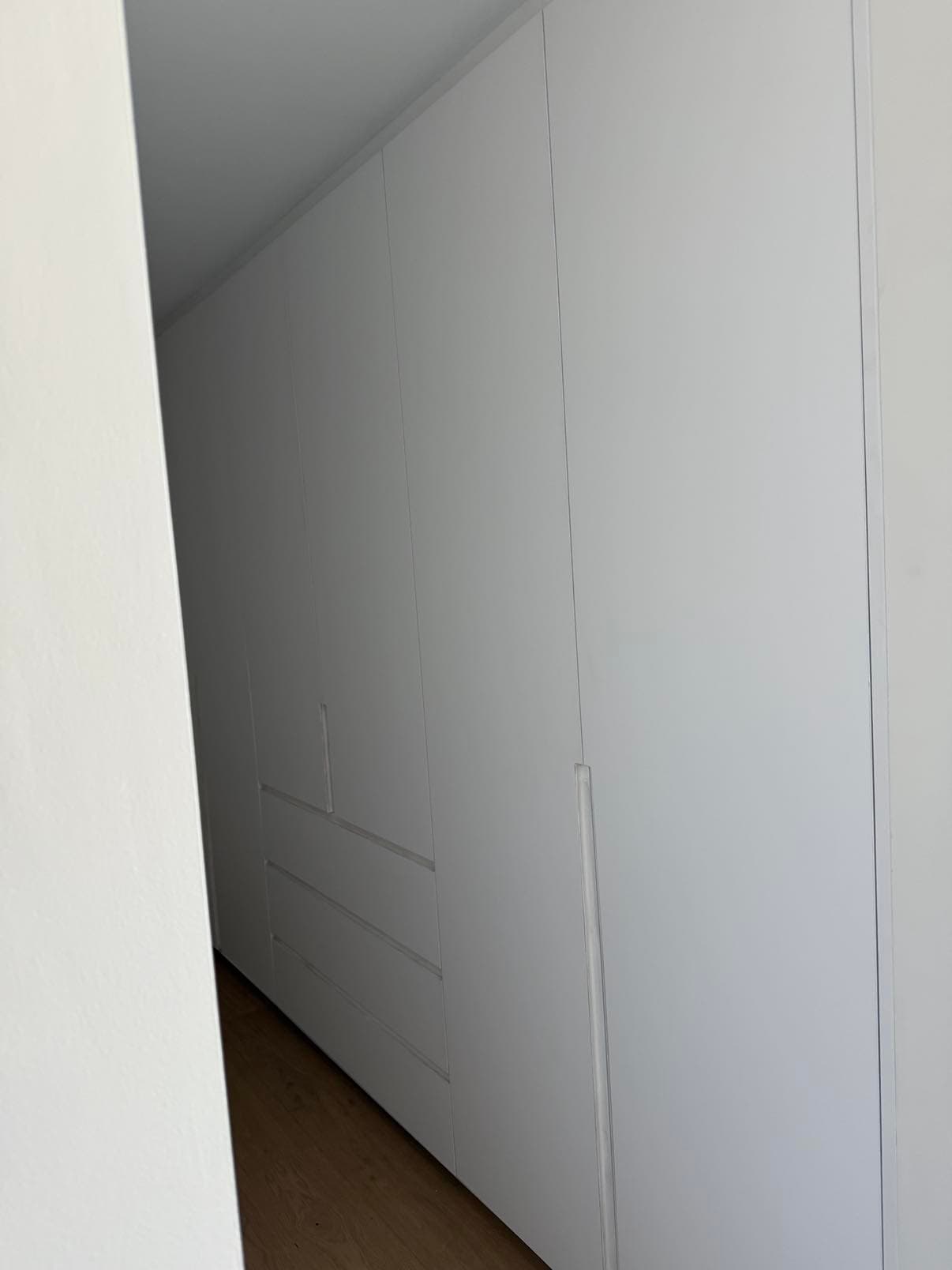 Alignement de placards blancs toute hauteur sans poignées, recoupés en biais pour suivre la pente d'un toit.
