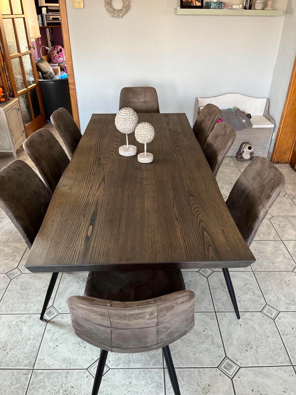 Grande table de salle à manger au plateau en frêne teinté brun, posée sur de fins pieds métalliques noirs et entourée de chaises.