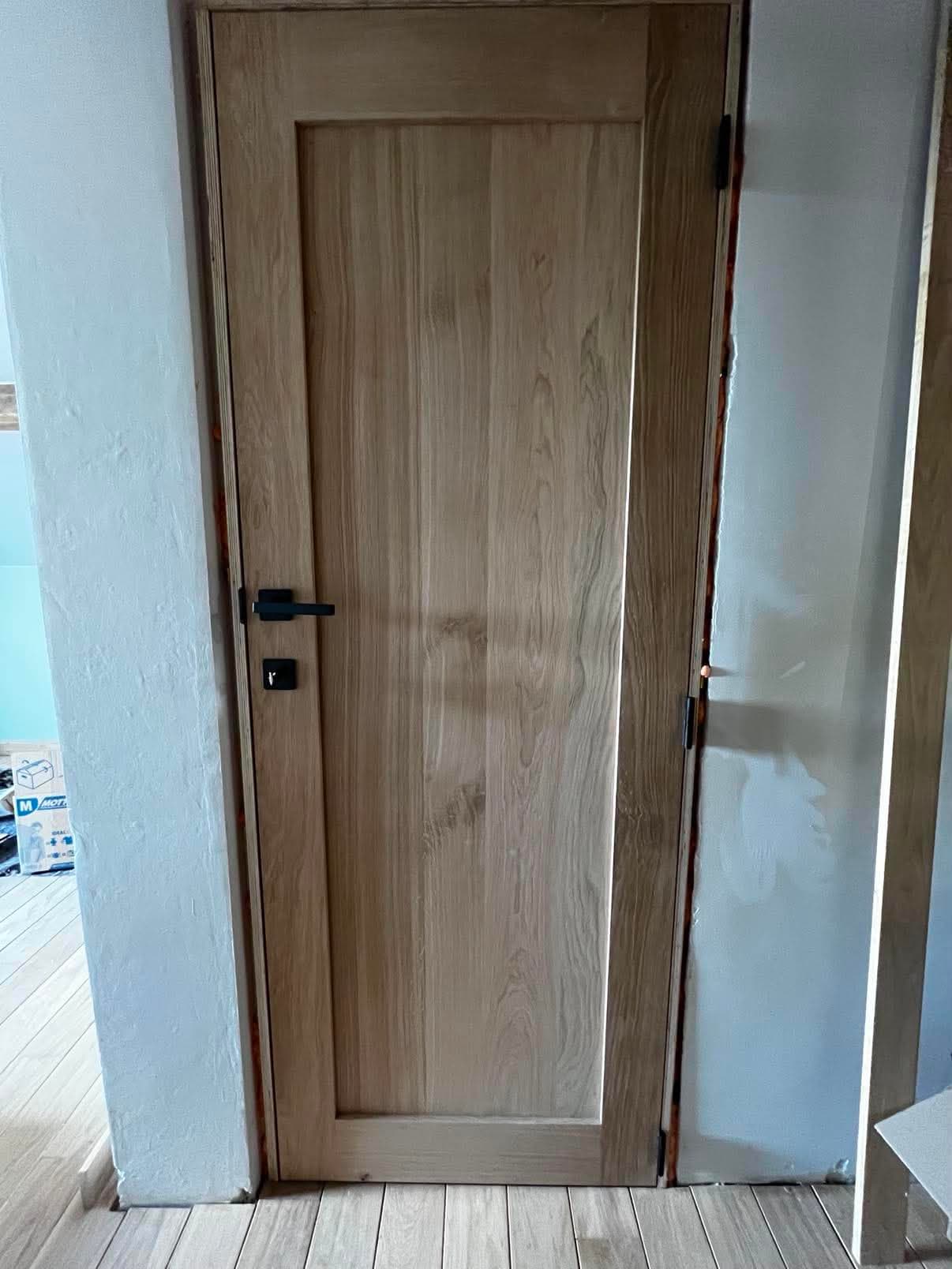 Porte intérieure en chêne massif à grand panneau, poignée et charnières noires, dans un logement en chantier.