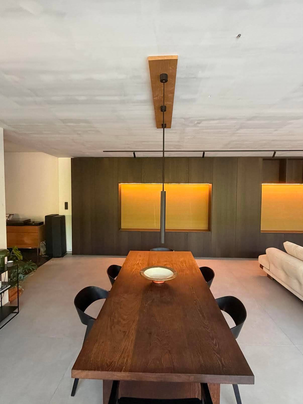 Table de repas en chêne teinté dans un séjour ouvert, face à un mur d'armoires foncé percé de niches rétroéclairées.