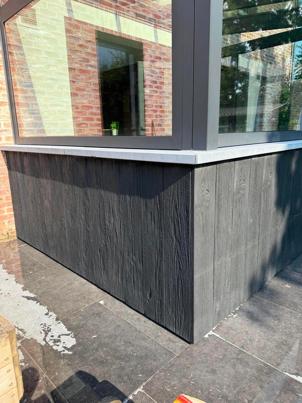 Soubassement de véranda habillé de planches de bois brûlé noir, sous des châssis en aluminium gris.