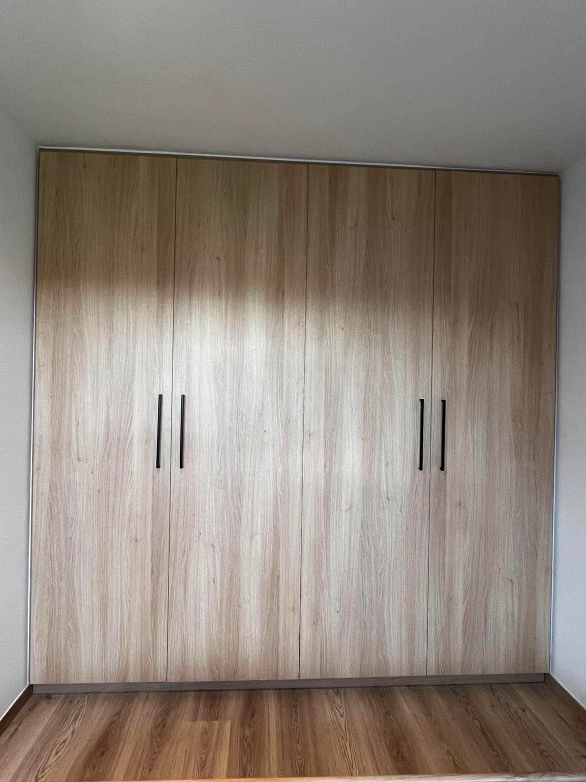 Armoire quatre portes toute hauteur en panneau décor chêne clair, ajustée entre deux murs, avec poignées bâton noires.