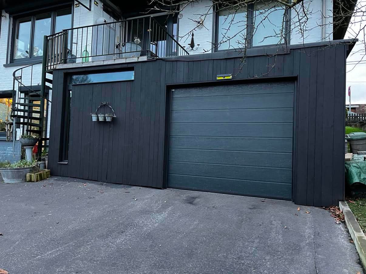 Garage accolé à une maison, habillé d'un bardage en lames de bois verticales teintées noir, avec porte sectionnelle.
