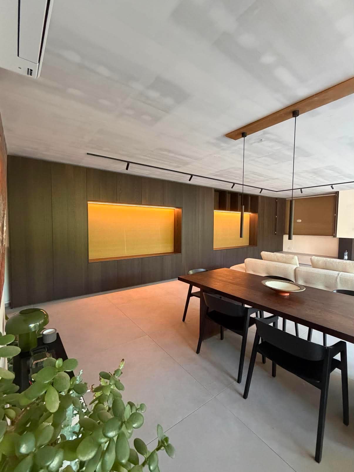 Séjour ouvert bordé de rangements toute hauteur en placage foncé, avec niches rétroéclairées et table de repas en bois.