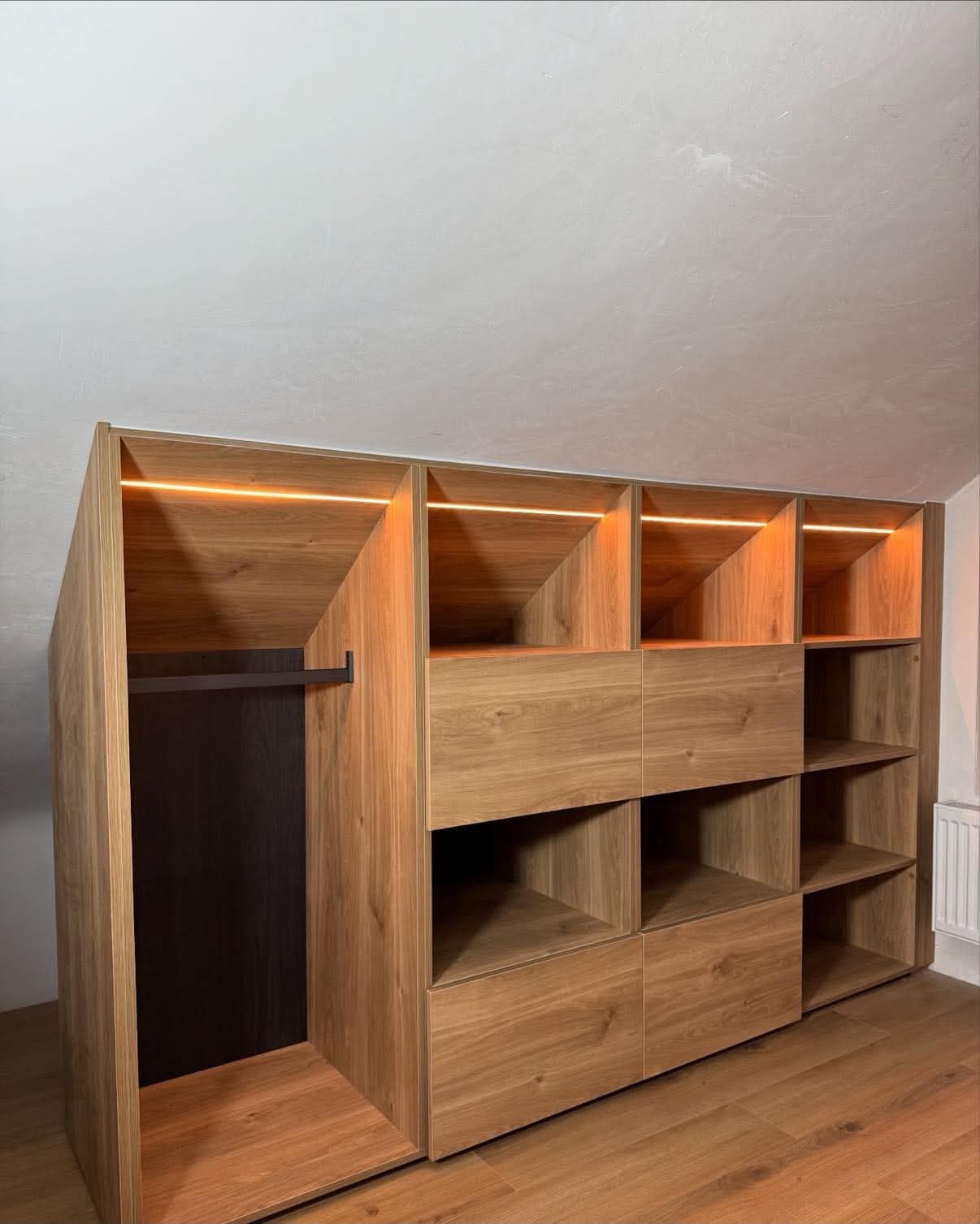 Meuble de rangement sous pente en panneau décor chêne, composé d'une penderie, de tiroirs et de niches éclairées par un ruban LED.