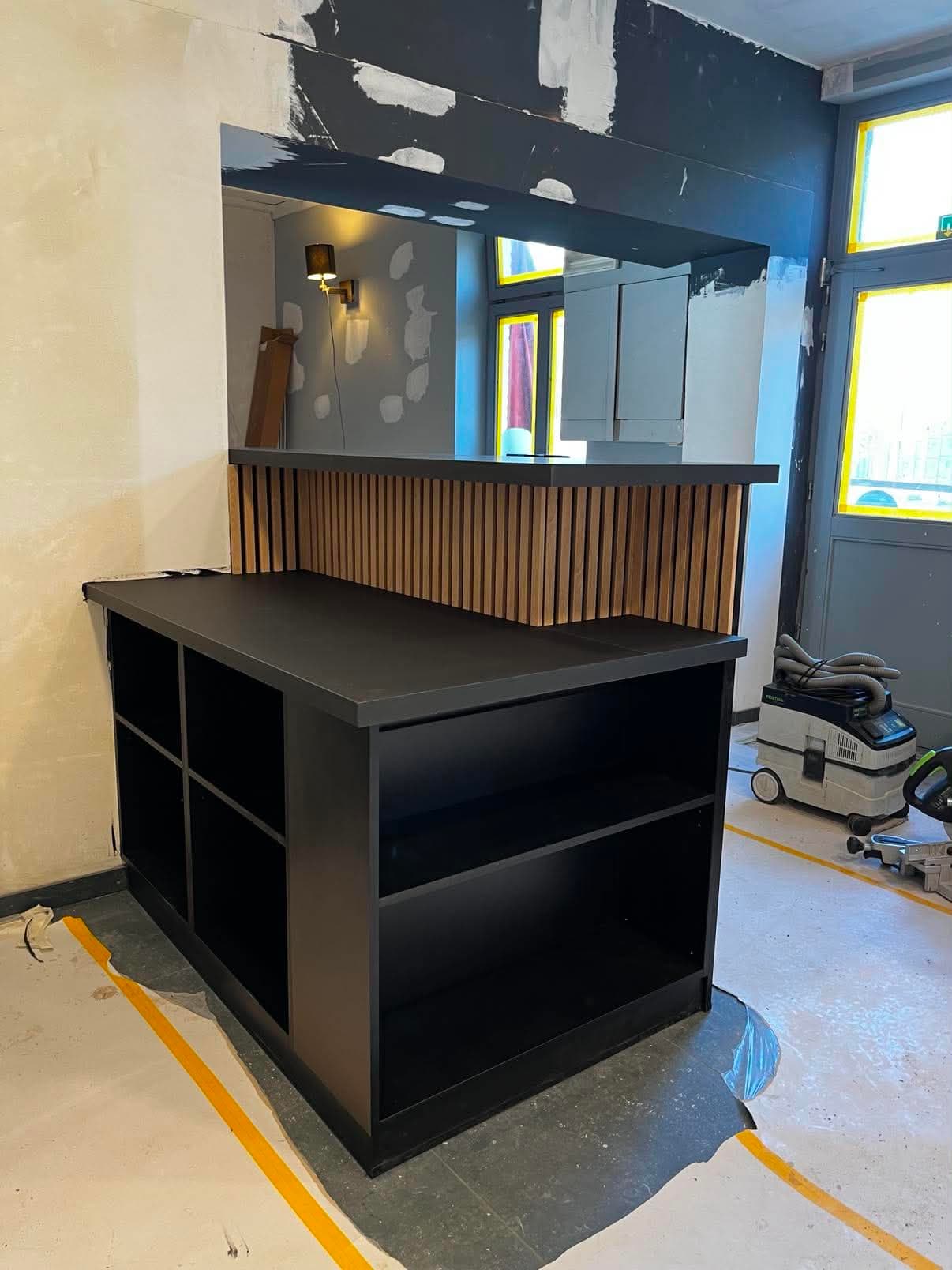 Comptoir de service en L à plan noir mat, avec un dosseret en tasseaux de chêne, dans un local en chantier.