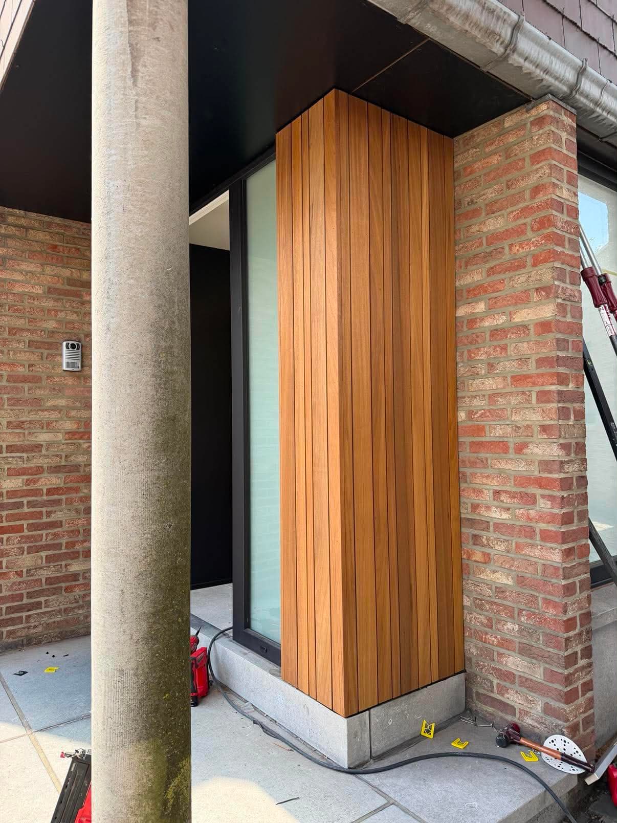 Pilier d'entrée de maison habillé d'un bardage en lames de bois verticales, entre un mur de briques et une baie vitrée.